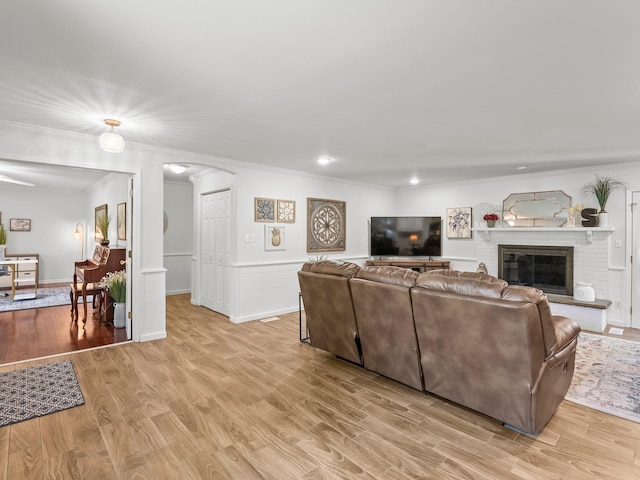 living room featuring arched walkways, crown molding, light wood-style flooring, a wainscoted wall, and a brick fireplace