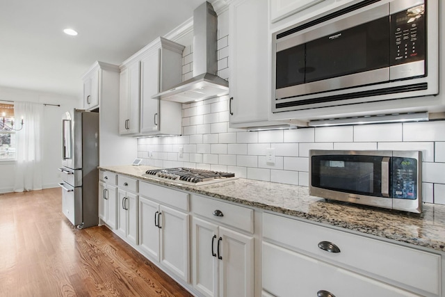 kitchen with stainless steel appliances, wall chimney exhaust hood, white cabinets, tasteful backsplash, and light wood-type flooring