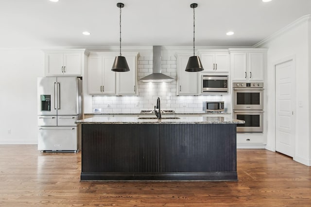 kitchen with appliances with stainless steel finishes, wall chimney exhaust hood, a center island with sink, ornamental molding, and backsplash
