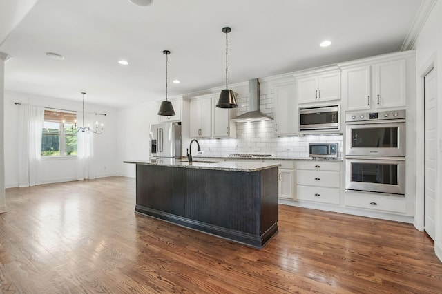 kitchen with wall chimney range hood, appliances with stainless steel finishes, a sink, a chandelier, and light stone counters