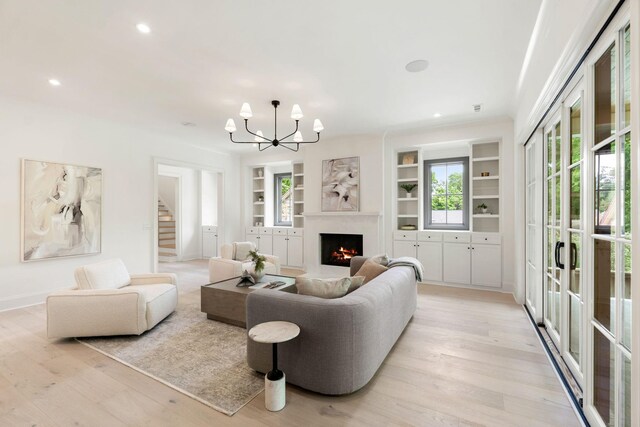 living room featuring stairs, a chandelier, built in shelves, a warm lit fireplace, and light wood-type flooring