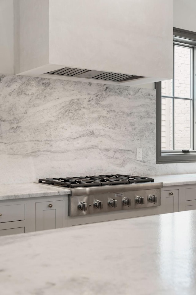 kitchen view of stainless steel gas stovetop, white cabinetry, light stone countertops, premium range hood, and tasteful backsplash