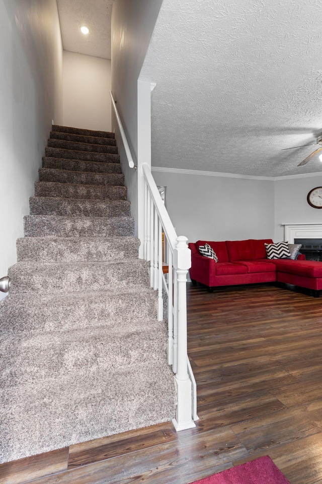 staircase with a ceiling fan, wood finished floors, a textured ceiling, ornamental molding, and a fireplace