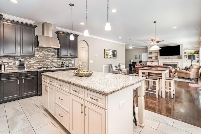 kitchen featuring wall chimney range hood, ceiling fan, ornamental molding, arched walkways, and a fireplace