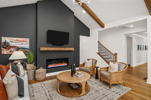 living room featuring wood finished floors, beamed ceiling, high vaulted ceiling, a glass covered fireplace, and stairs