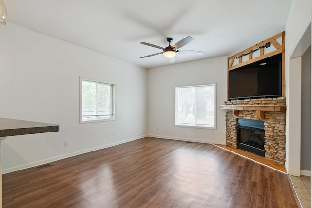 unfurnished living room with a ceiling fan, wood finished floors, a stone fireplace, and baseboards