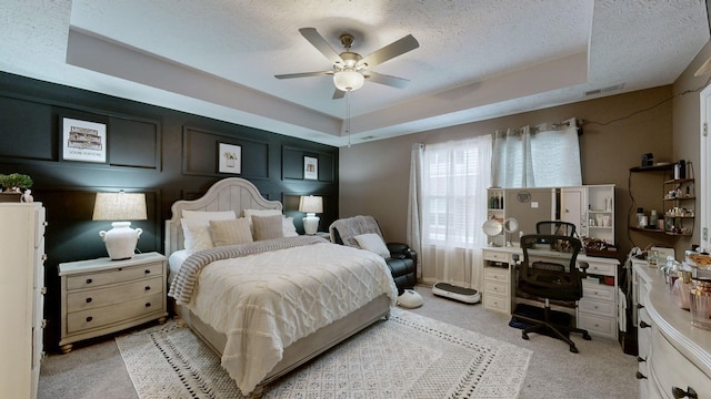 bedroom with a textured ceiling, light carpet, a tray ceiling, ceiling fan, and an office area