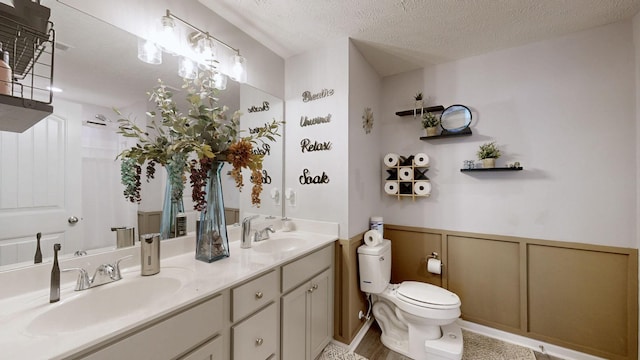 full bathroom featuring toilet, double vanity, wainscoting, a textured ceiling, and a decorative wall