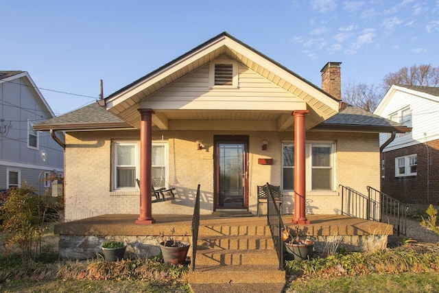 bungalow-style house featuring a porch, a shingled roof, a chimney, and brick siding