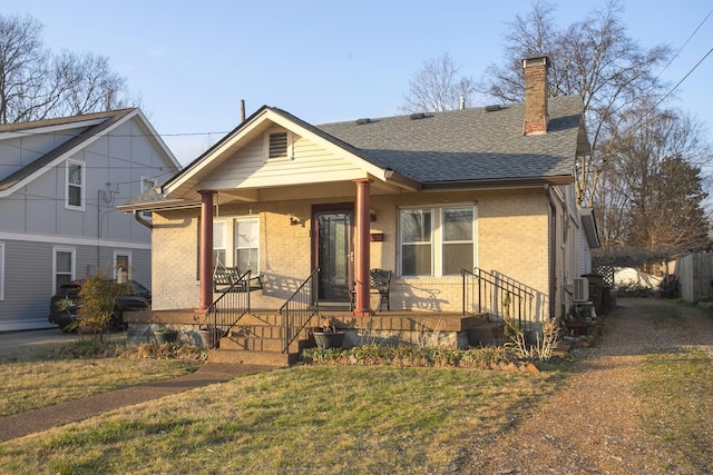 bungalow-style home with covered porch, a chimney, brick siding, roof with shingles, and a front lawn