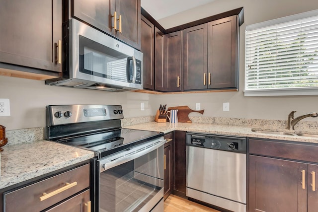kitchen with stainless steel appliances, dark brown cabinetry, a sink, light stone counters, and light wood finished floors