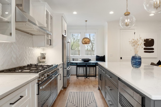 kitchen featuring high end appliances, wall chimney exhaust hood, light wood finished floors, backsplash, and white cabinetry