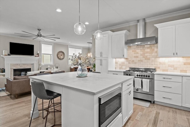 kitchen with stainless steel appliances, wall chimney exhaust hood, a fireplace, a kitchen island, and light wood-style flooring