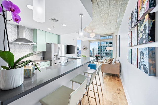 kitchen featuring wall chimney exhaust hood, stainless steel refrigerator with ice dispenser, light wood finished floors, baseboards, and a breakfast bar area