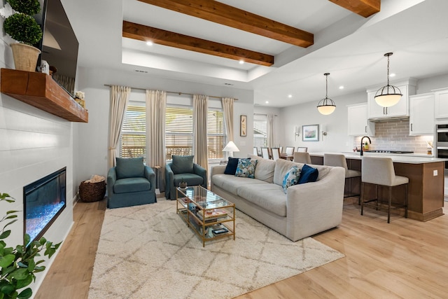 living area featuring light wood-type flooring, recessed lighting, beamed ceiling, and a glass covered fireplace