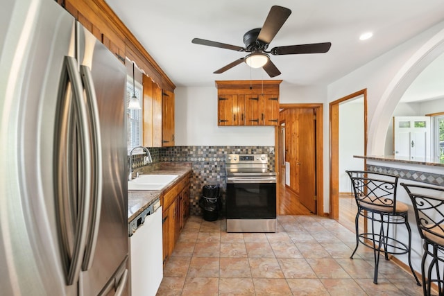 kitchen with appliances with stainless steel finishes, a sink, decorative backsplash, brown cabinetry, and ceiling fan
