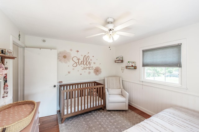 bedroom featuring wood finished floors, wainscoting, a nursery area, and ceiling fan