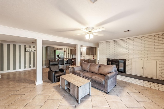 living room featuring a ceiling fan, a brick fireplace, light tile patterned floors, crown molding, and baseboards