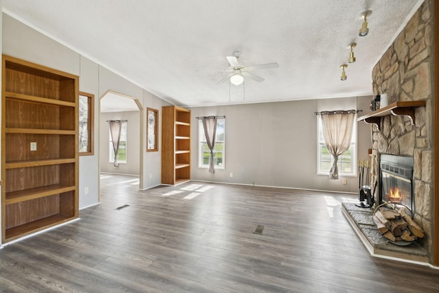 unfurnished living room with a textured ceiling, a ceiling fan, a stone fireplace, ornamental molding, and dark wood finished floors
