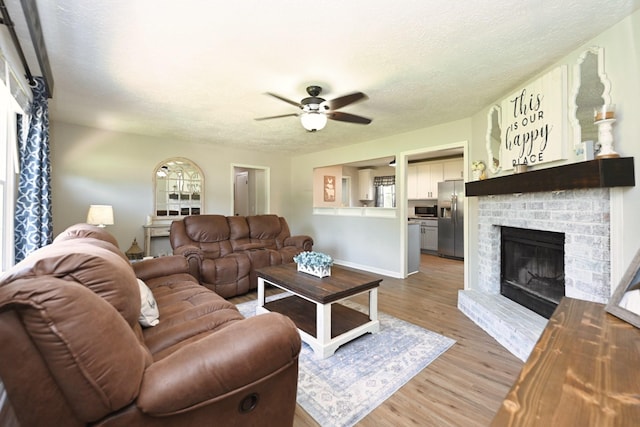 living area featuring a brick fireplace, a ceiling fan, light wood-style flooring, baseboards, and a textured ceiling