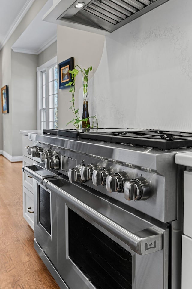 kitchen view of range with two ovens, wall chimney range hood, crown molding, light wood-type flooring, and baseboards