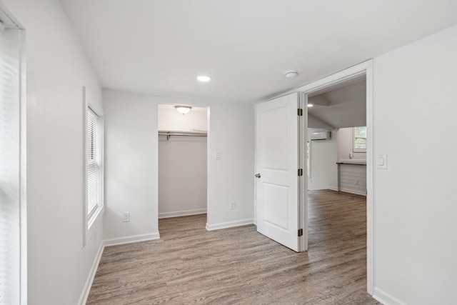 unfurnished bedroom with light wood-style flooring, a wall unit AC, a walk in closet, baseboards, and multiple windows