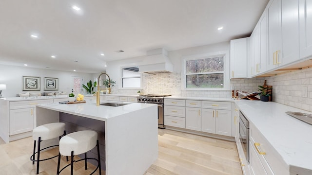 kitchen with stainless steel appliances, a sink, custom range hood, decorative backsplash, and a center island with sink