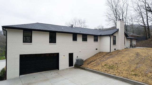back of house featuring a garage, roof with shingles, concrete driveway, a chimney, and brick siding