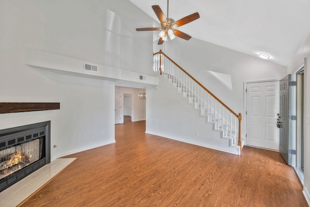 unfurnished living room with high vaulted ceiling, stairway, ceiling fan, a fireplace with flush hearth, and wood finished floors