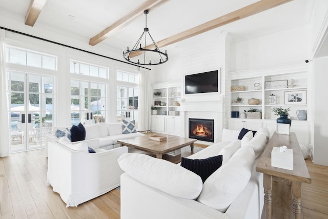 living room with beamed ceiling, a chandelier, a glass covered fireplace, light wood-style floors, and built in shelves