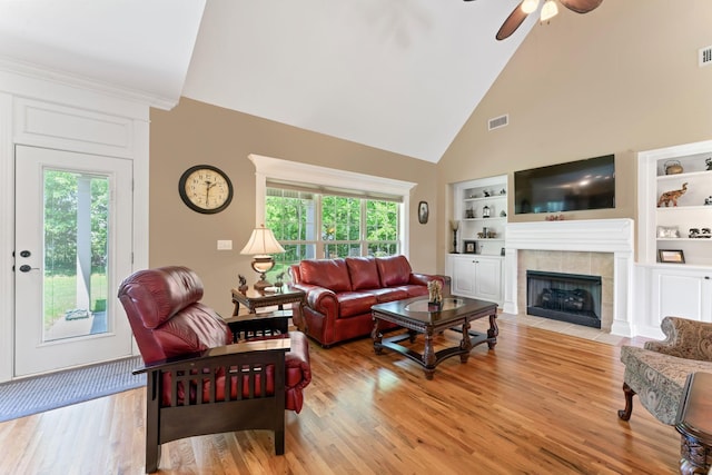 living room with a ceiling fan, a tile fireplace, light wood-style flooring, high vaulted ceiling, and built in shelves