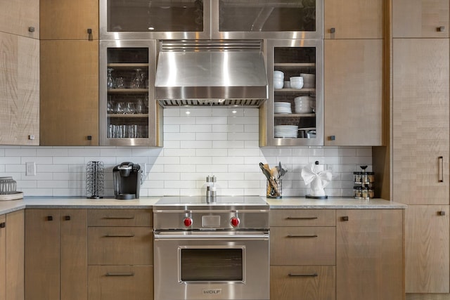 kitchen featuring luxury range, wall chimney range hood, tasteful backsplash, light countertops, and glass insert cabinets