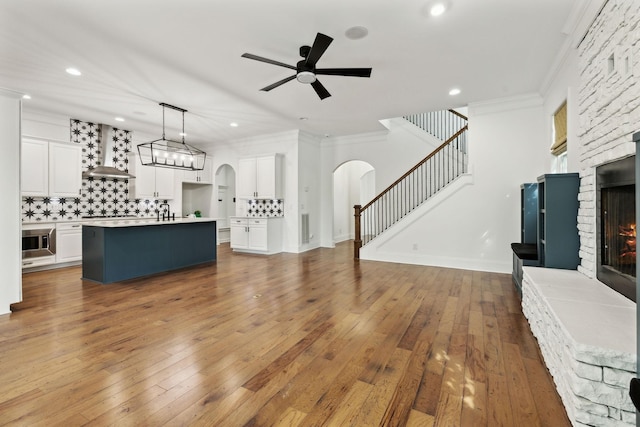 unfurnished living room featuring arched walkways, stairs, ceiling fan, a warm lit fireplace, and ornamental molding