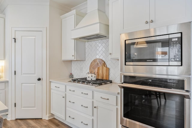 kitchen with appliances with stainless steel finishes, custom exhaust hood, backsplash, light wood-style floors, and white cabinetry