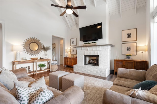 living room with high vaulted ceiling, ceiling fan, wood finished floors, a brick fireplace, and beamed ceiling