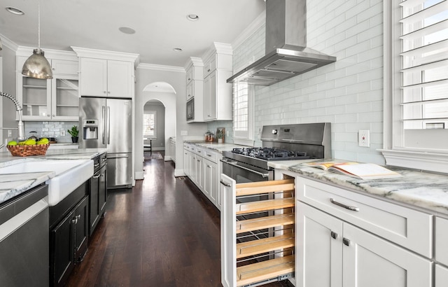 kitchen with stainless steel appliances, arched walkways, wall chimney range hood, decorative backsplash, and ornamental molding