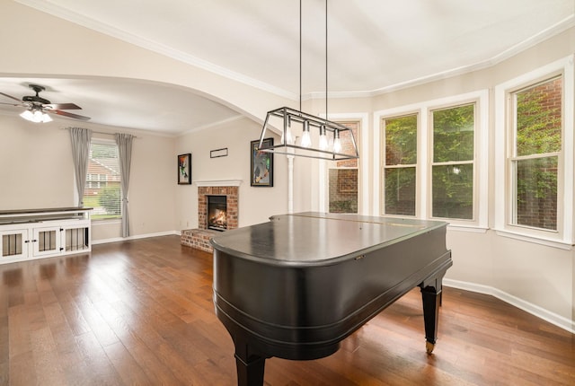game room featuring dark wood-style flooring, baseboards, a ceiling fan, a brick fireplace, and crown molding