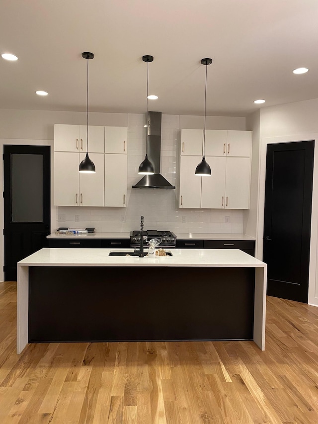 kitchen featuring wall chimney exhaust hood, a sink, an island with sink, white cabinets, and light countertops