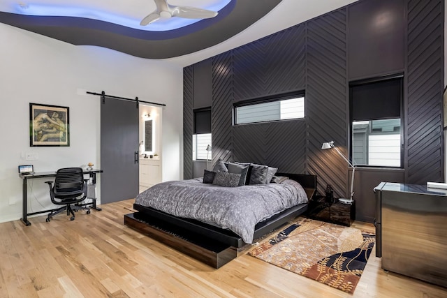 bedroom featuring a barn door, light wood-type flooring, a desk, ceiling fan, and connected bathroom