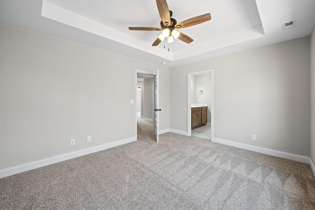 unfurnished bedroom with a raised ceiling, light carpet, baseboards, ensuite bathroom, and a ceiling fan
