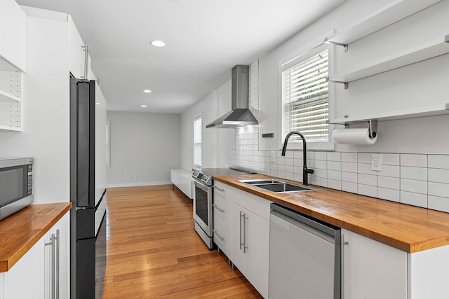 kitchen featuring wooden counters, open shelves, stainless steel appliances, wall chimney exhaust hood, and a sink
