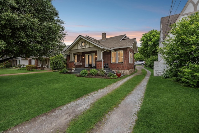 view of front facade with driveway, brick siding, a chimney, a lawn, and covered porch