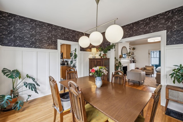 dining area with wallpapered walls, light wood-style flooring, wainscoting, a fireplace, and a decorative wall