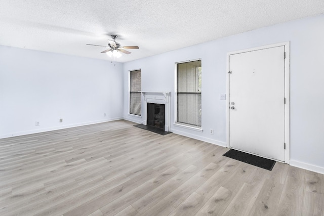 unfurnished living room with ceiling fan, light wood-style floors, a fireplace with raised hearth, a textured ceiling, and baseboards
