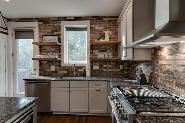 kitchen featuring open shelves, wall chimney range hood, appliances with stainless steel finishes, a sink, and dark stone counters