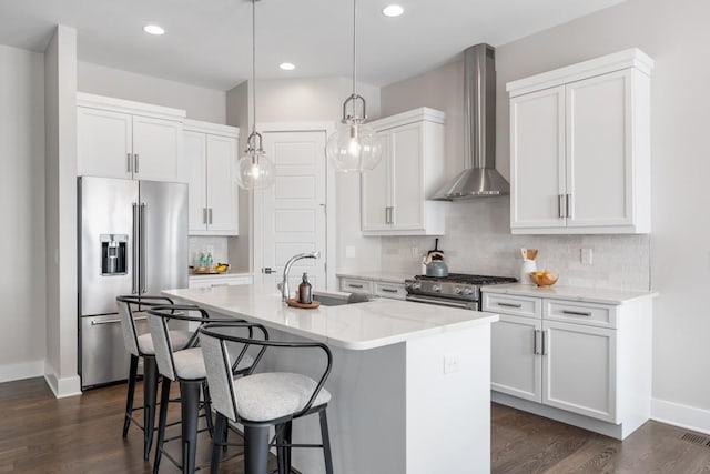 kitchen featuring high quality appliances, wall chimney range hood, a sink, baseboards, and a center island with sink