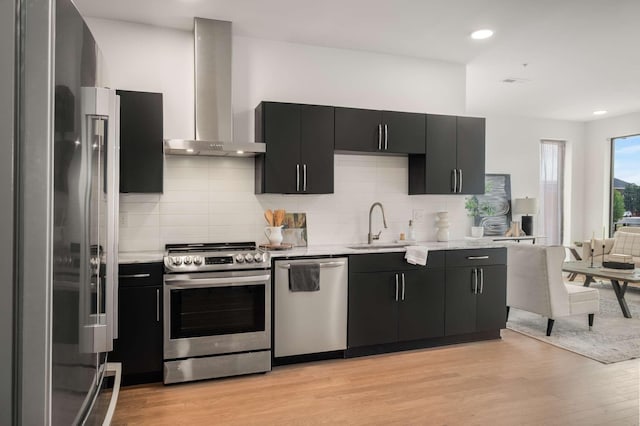 kitchen with appliances with stainless steel finishes, wall chimney range hood, dark cabinetry, a sink, and light wood-type flooring