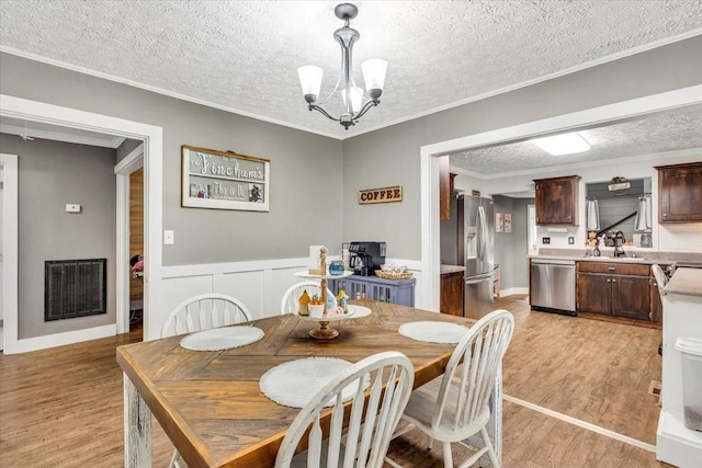 dining space featuring a textured ceiling, light wood-style floors, a chandelier, a wainscoted wall, and ornamental molding