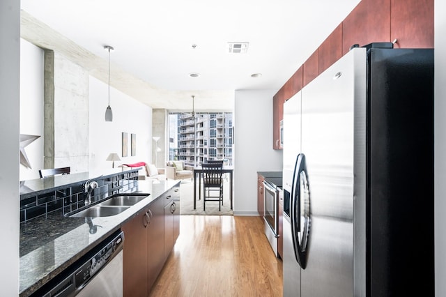 kitchen with appliances with stainless steel finishes, a sink, light wood-type flooring, decorative light fixtures, and dark stone counters