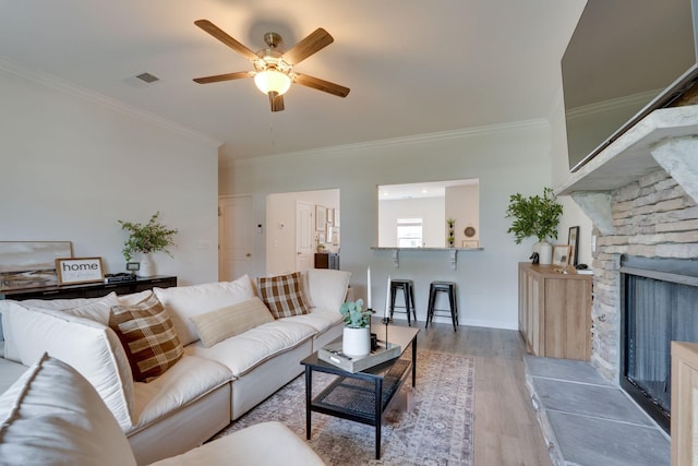 living room featuring a ceiling fan, light wood-type flooring, crown molding, and a stone fireplace